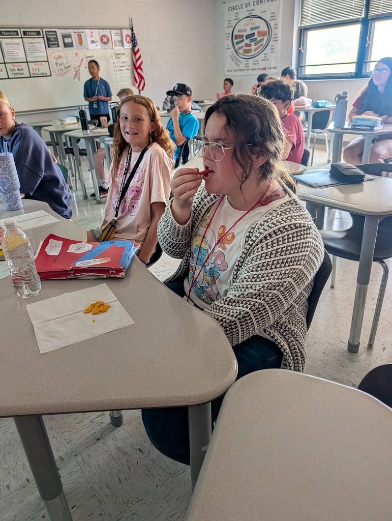 A classroom with desks, students, one eating a snack with utensils, a flag, and a teacher.