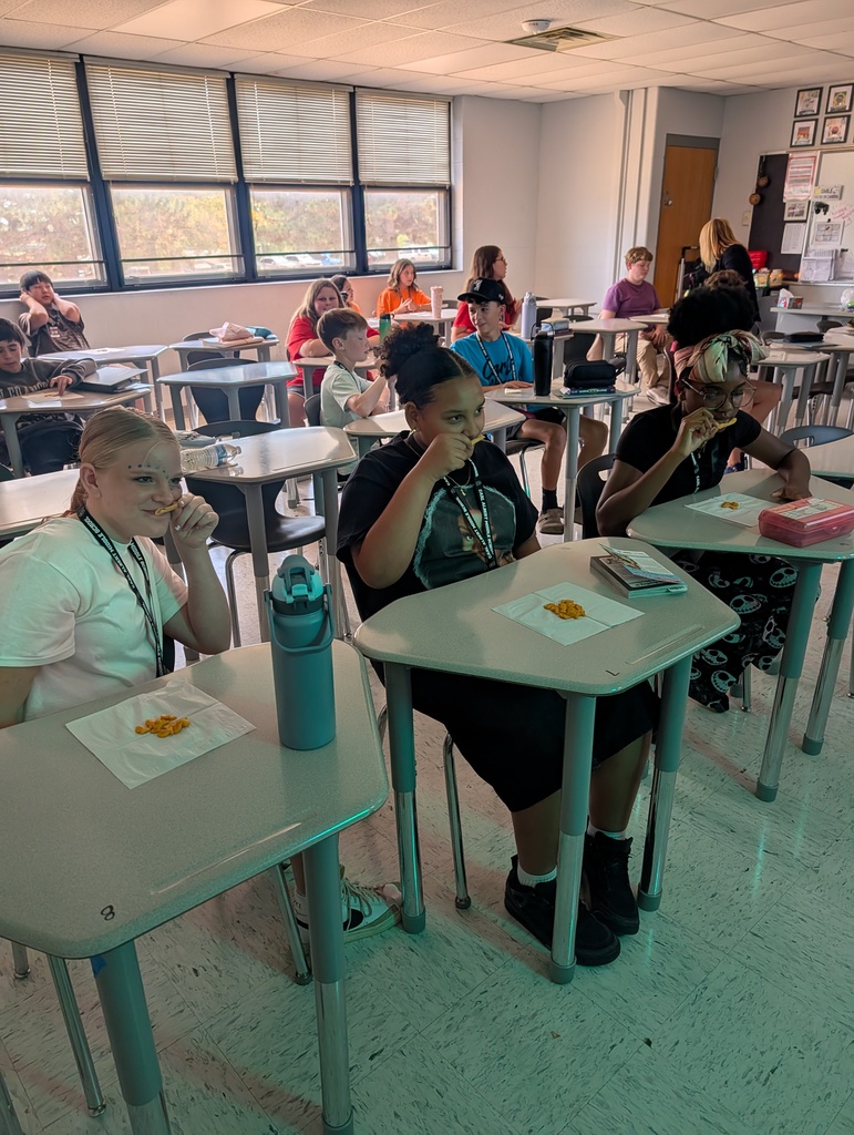 Students sitting at desks in a classroom, with one girl eating chips and another wearing a headpiece.