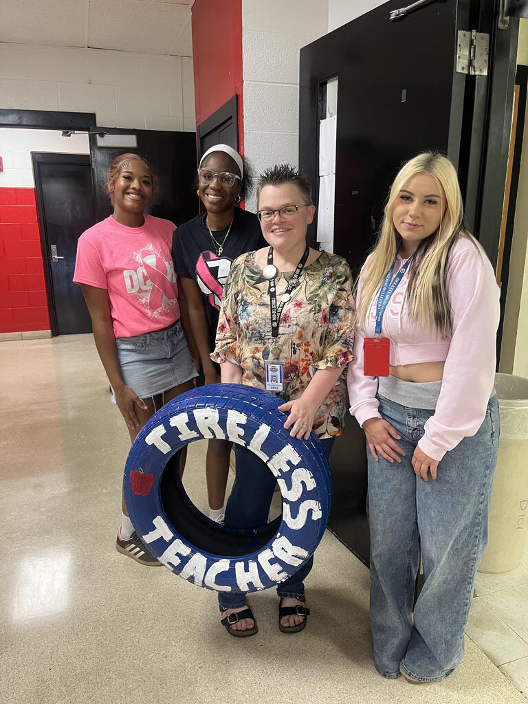 Four women pose for a photo in a hallway. One holds a tire sign. Two wear sneakers; one wears sandals. Background includes doors and a trash can.