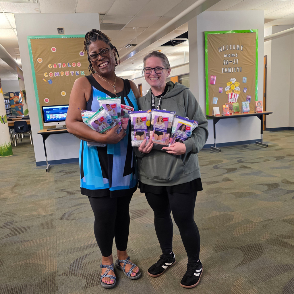 A picture of 2 women standing in the MCMS library holding multiple packs of underwear with huge smiles on their faces.