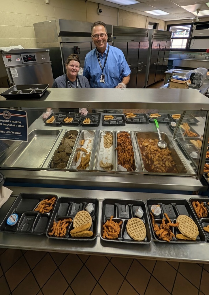 Two adults standing behind a counter with trays of food.