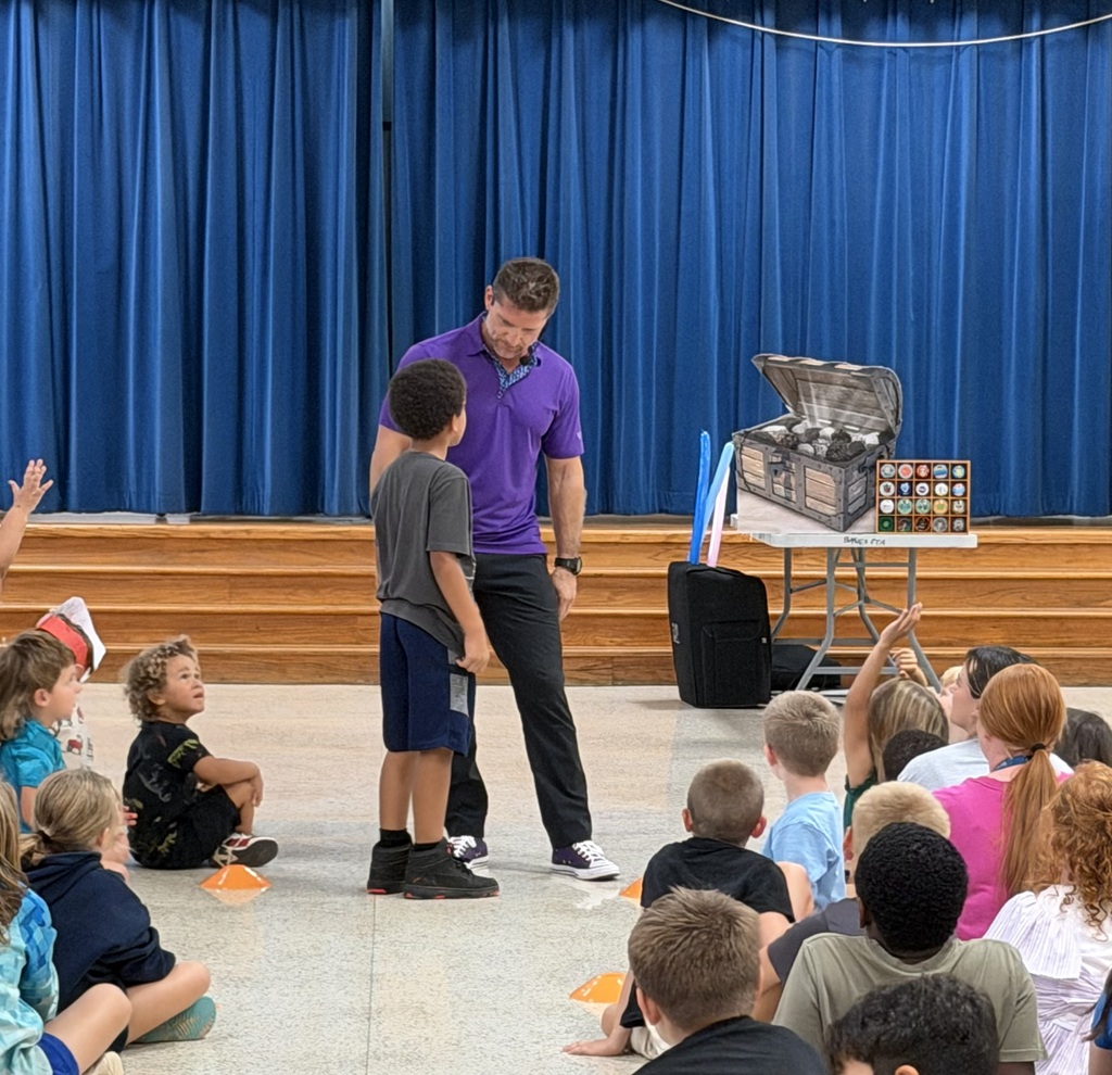An indoor area with blue curtains. A man and child are standing before a seated audience of children.