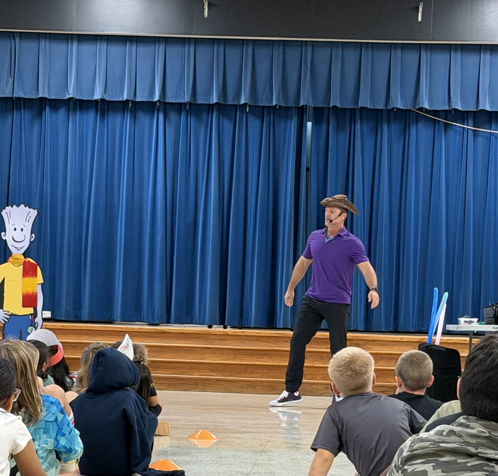 An audience watching a person on stage wearing a hat and purple shirt. Behind the person, blue curtains.