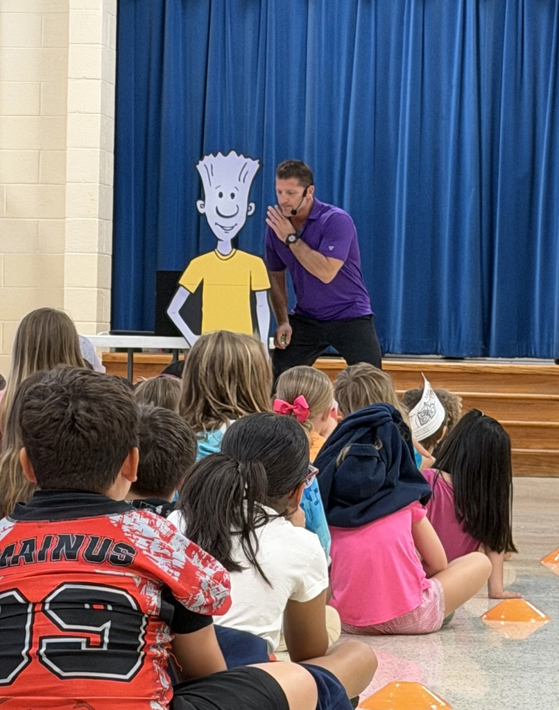 A person wearing a microphone stands in front of a large cartoon drawing. Children sit on the floor watching.