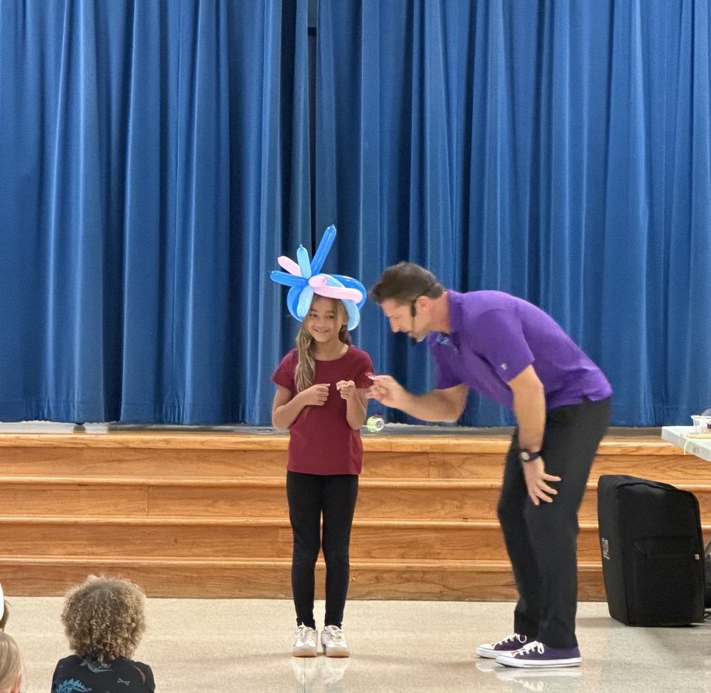 A person with a balloon hat stands next to a man in a purple shirt. They are on a stage with blue curtains and a wooden floor.