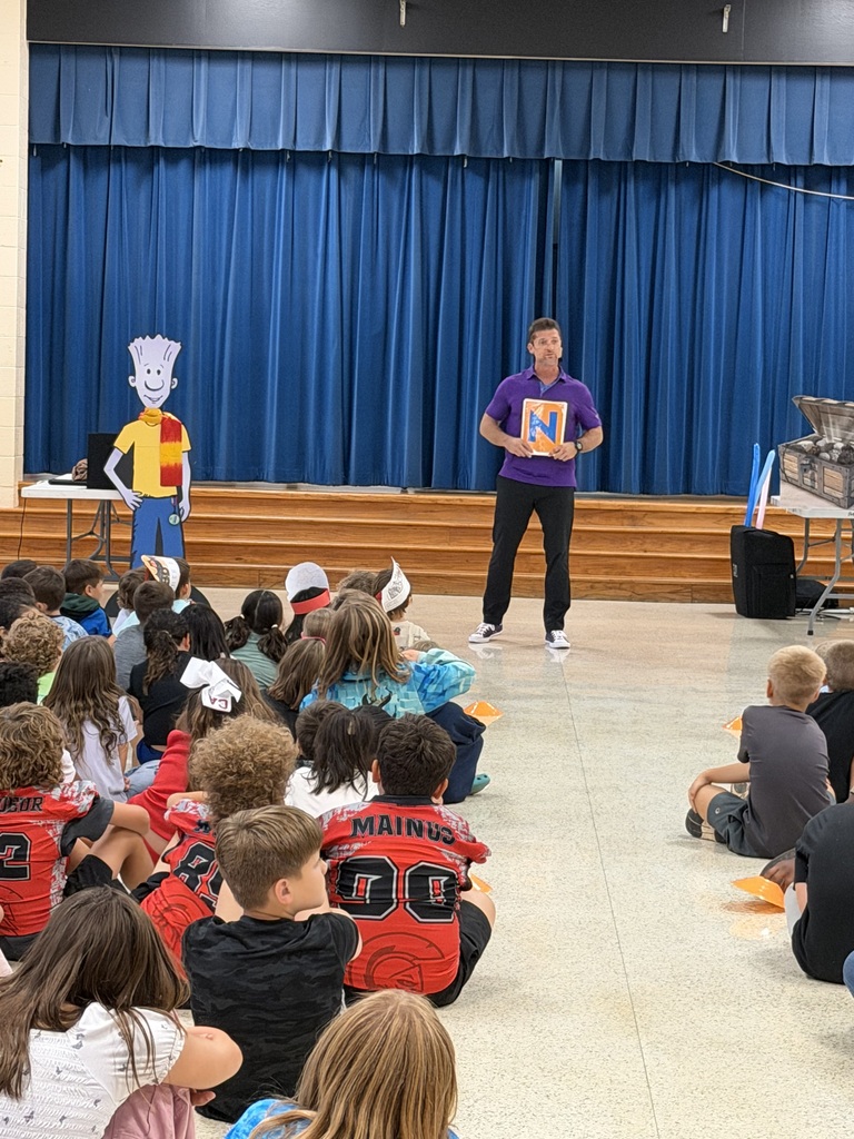 A man stands on stage with a puppet, addressing children in a classroom setting with blue curtains in the background.