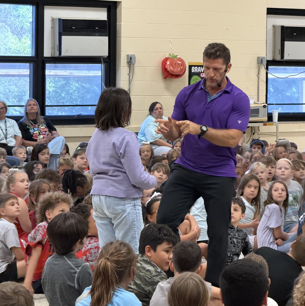 A man in purple stands before a classroom full of children. He gestures while speaking.