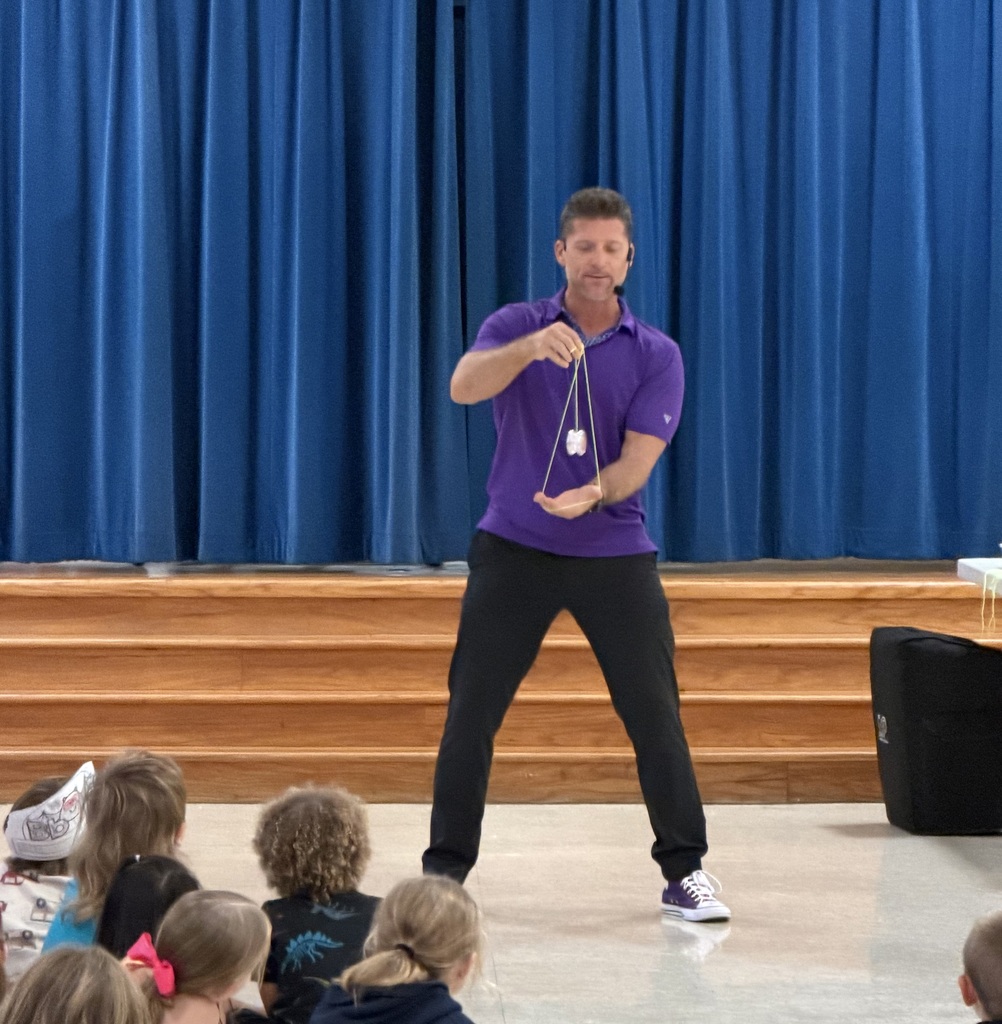 Man in purple shirt and black pants on stage, performing a magic trick with a string for a group of children.