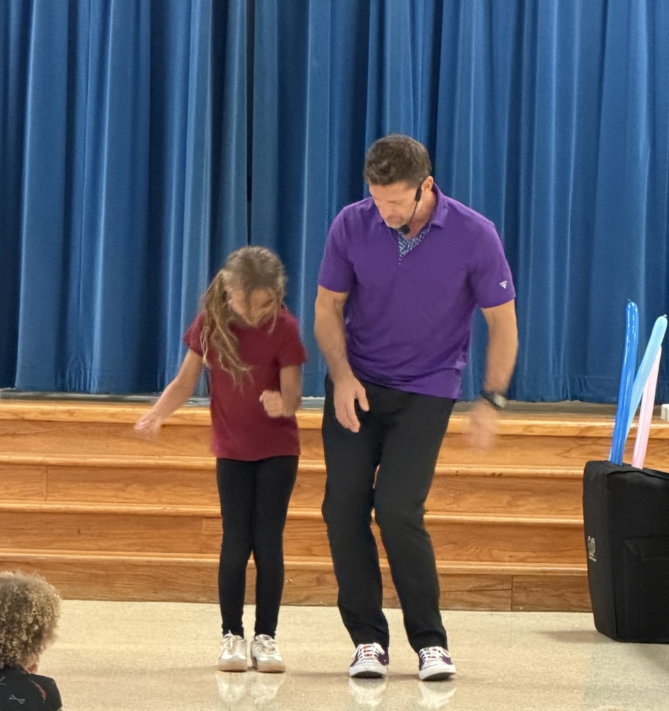 Man and girl in purple and red shirts on stage. Man leads girl in dance. Blue curtains in background.