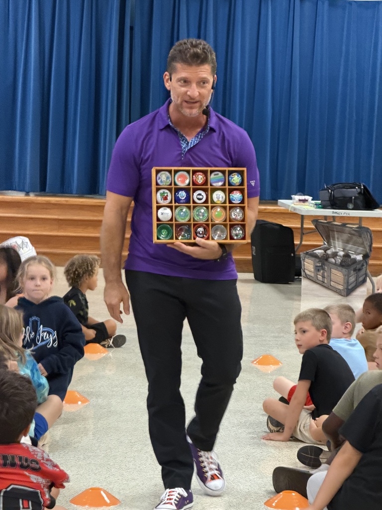 Man in purple shirt and black pants holds small box of yo-yos in front of seated children.