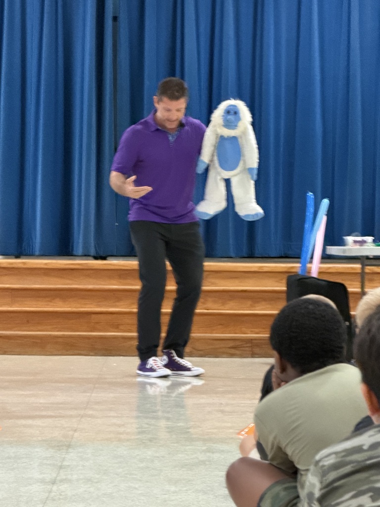 A man on stage, holding a white puppet, speaks to an audience. Behind him, a blue curtain and a table.