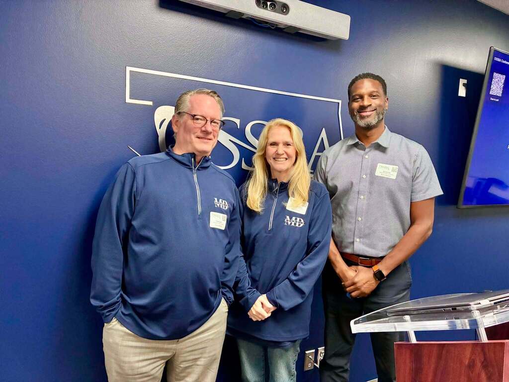 Three people in blue shirts stand together in front of a blue wall with an emblem.