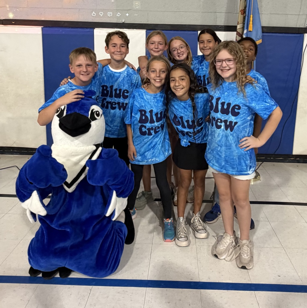Kids in blue shirts and shorts pose with a bluejay mascot in a gym.
