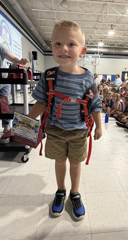 A young boy with a backpack smiles with a crowd of students seated.