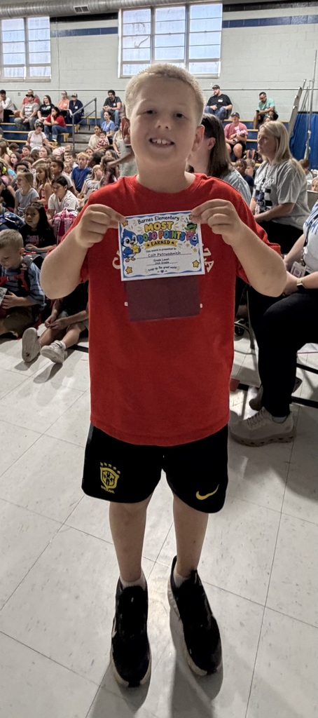 A young boy in a red shirt and black shorts holds up a certificate, smiling, with many people seated behind him in an indoor area.