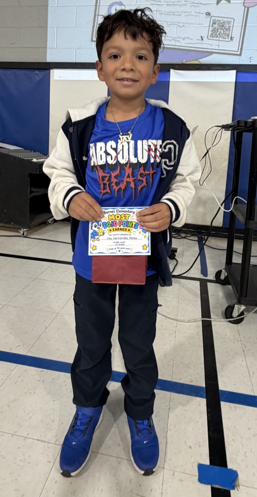 A young boy holds a certificate and smiles, wearing a blue shirt, black pants, and blue sneakers. He stands in a room with blue walls and blue lines on the floor.