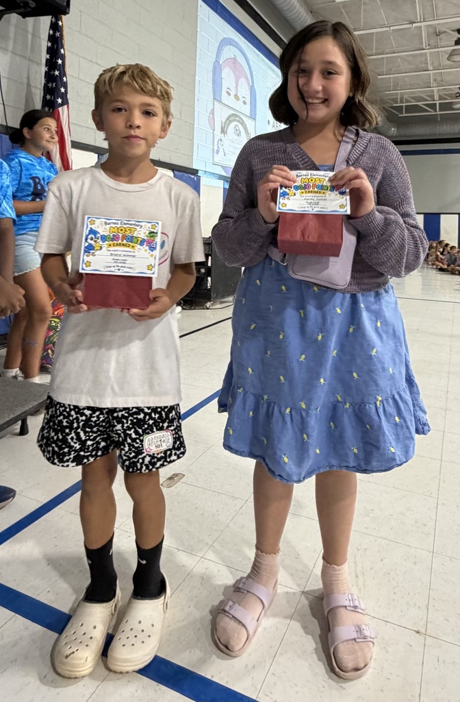 A boy and a girl hold certificates in an indoor setting with a flag and other people.