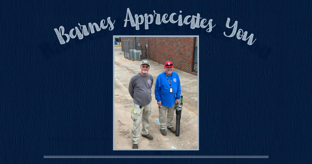 Two men in hats, standing on a dirt path, one holding a tool. A brick building is behind them.