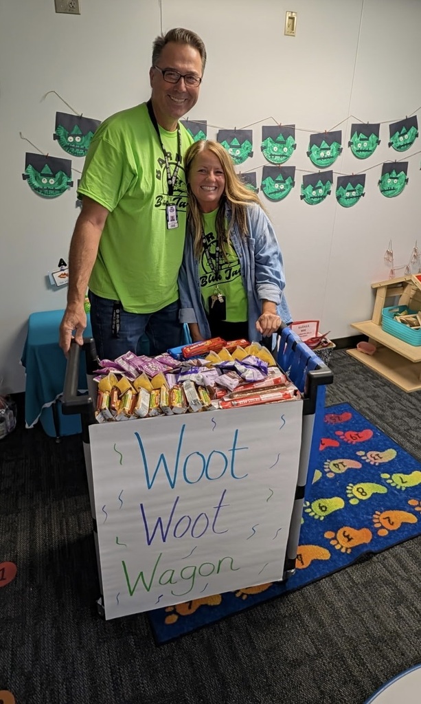 Two adults standing by a cart with a sign that reads "Woot Woot Wagon" in front of a wall.