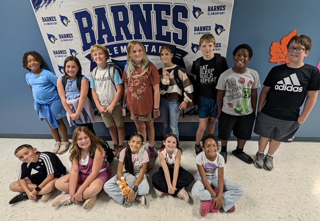 Several children in a group, standing and sitting on white floor, with a sign in the background reading "BARNES ELEMENTARY."