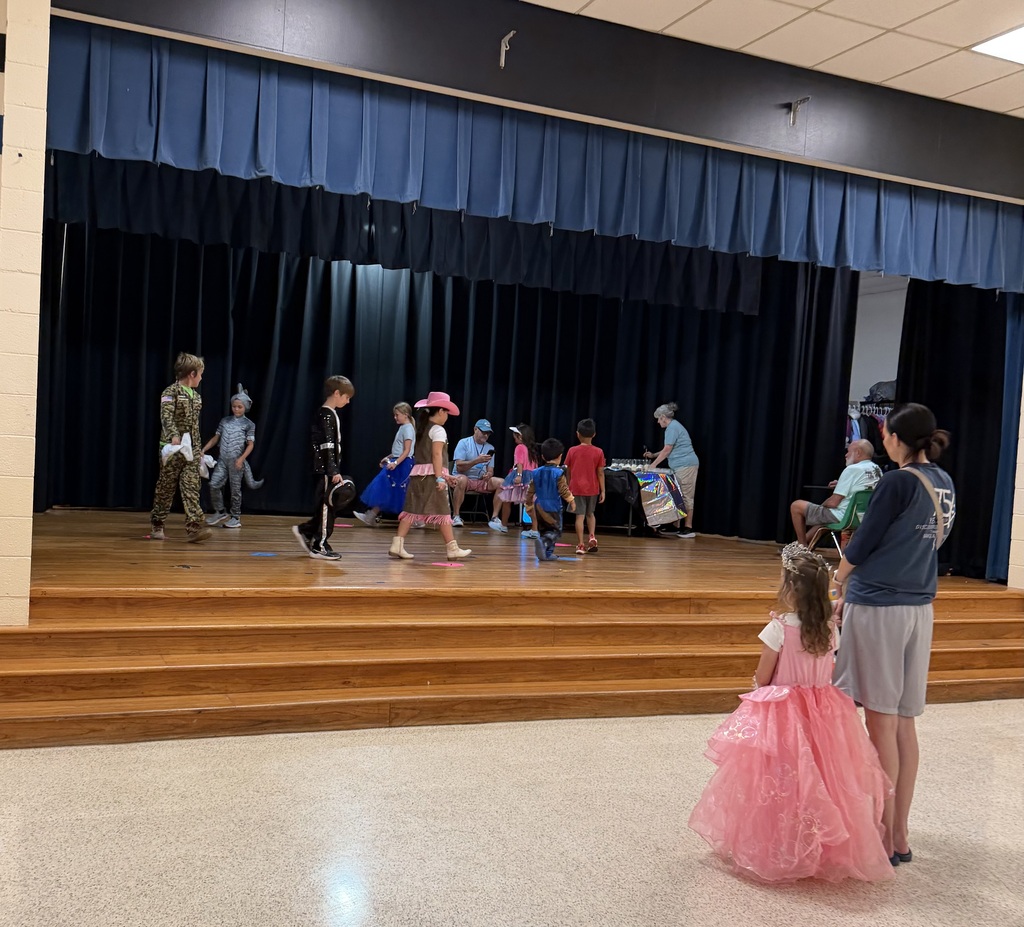 People stand on a stage in a theater. A young girl in a pink dress and another woman look on.
