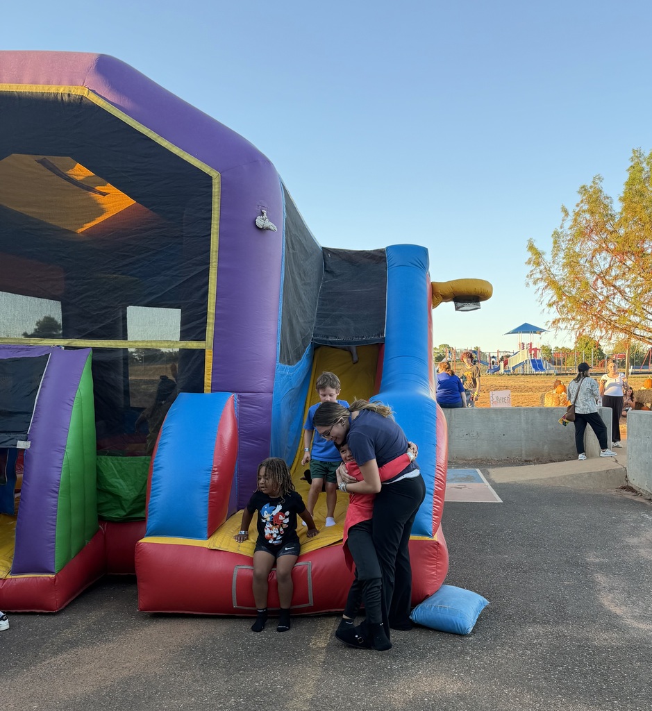 A group of children and an adult play in a colorful inflatable bounce house, set against a backdrop of a clear blue sky and trees.