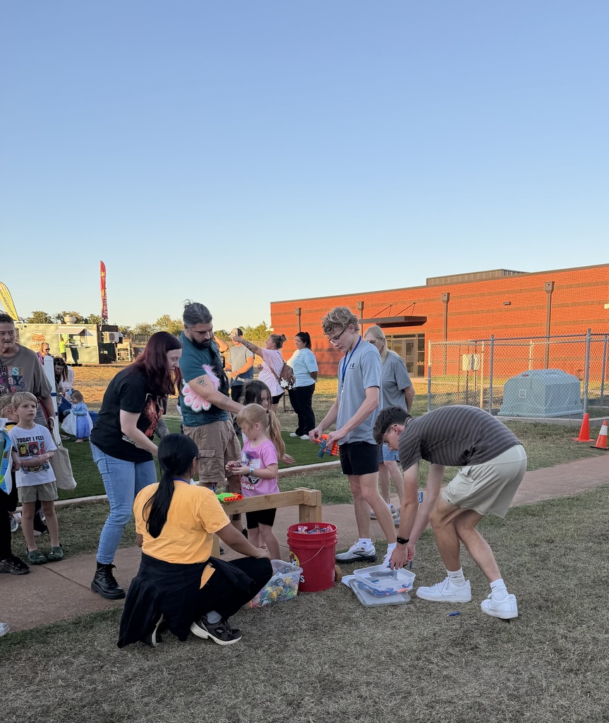 People engage in an outdoor activity. Some kneel and some stand. A red bucket is on the ground.