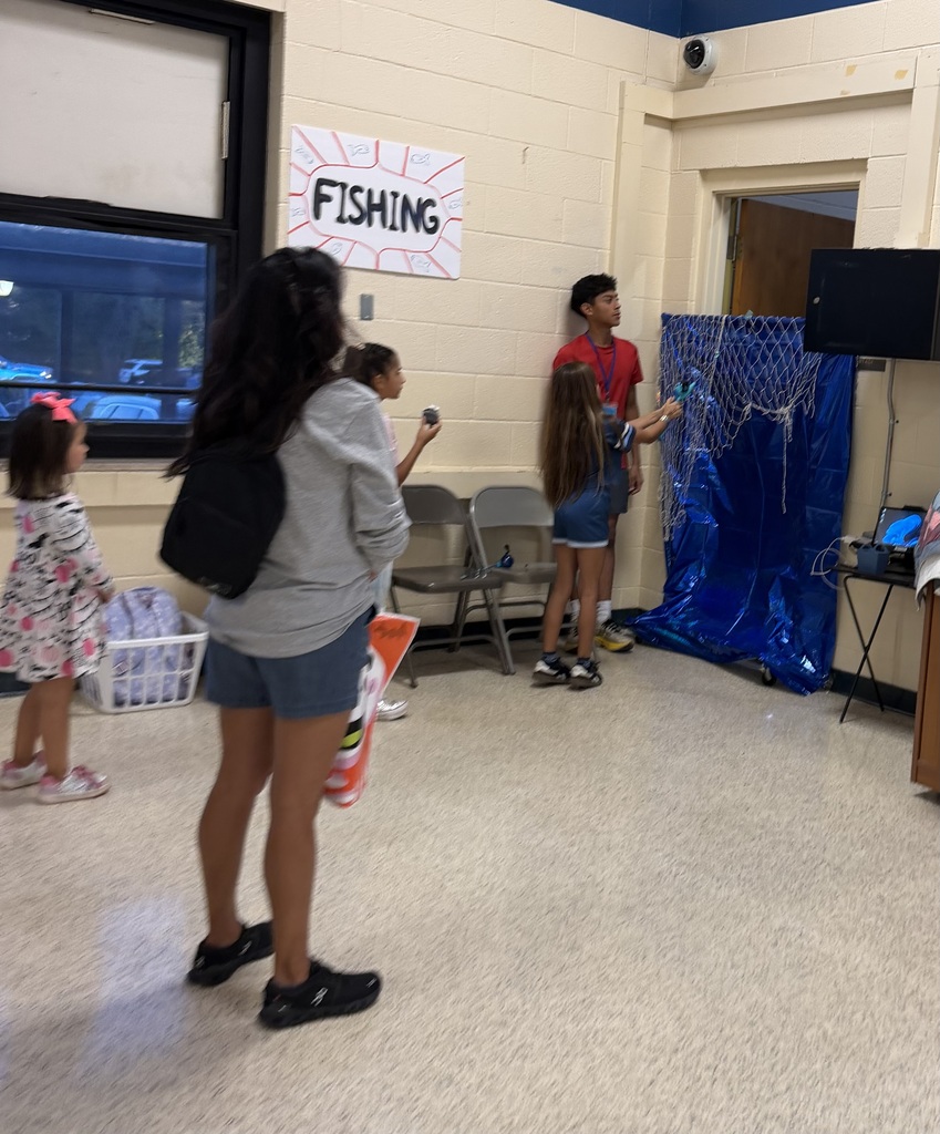 Children and adults stand in a room, two hold fish toys, and a blue tarp is on the wall.