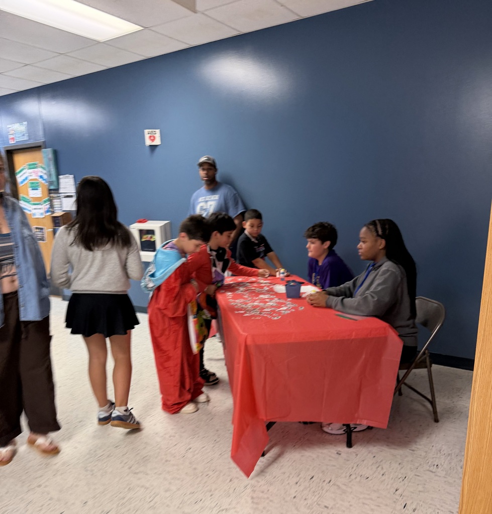 Multiple people gather around a table with a red cloth in a room with blue walls.