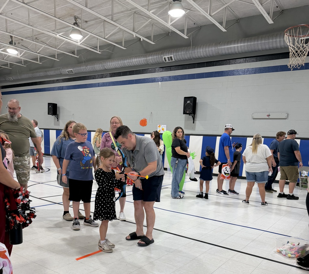 A group of adults and children interact inside a gymnasium. Some hold balloons. White flooring and blue wall.