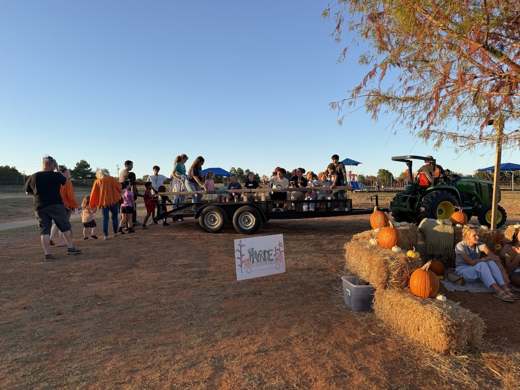 People gather around a tractor pulling a trailer with hay bales, pumpkins, and a sign.