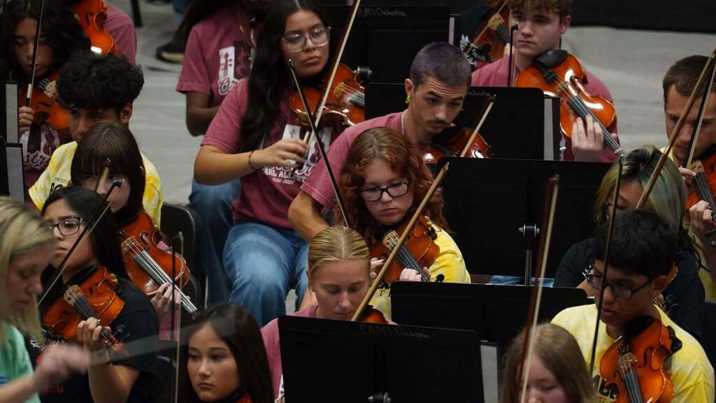A group of people in a room play violins and sit on chairs. They are holding violins and bows.