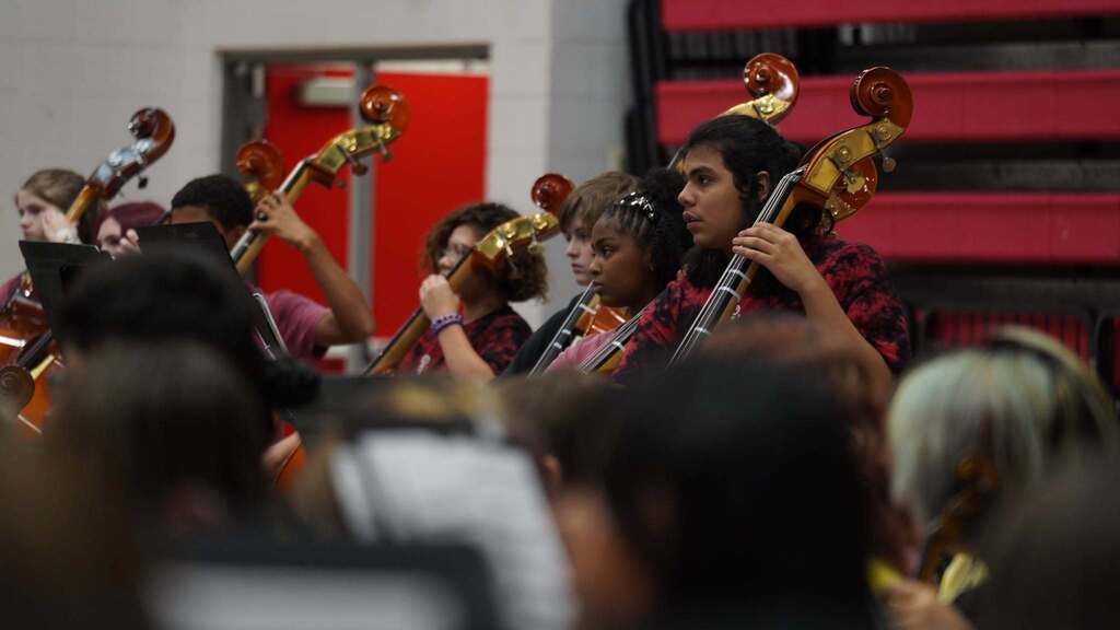 A group of people in a room play violins and sit on chairs. They are holding violins and bows.