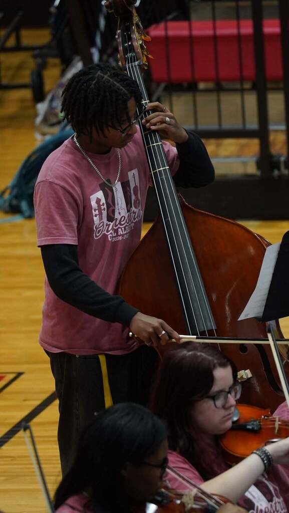 A student in a red shirt is standing playing the cello. 