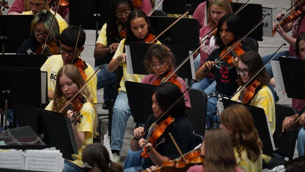 A group of people in a room play violins and sit on chairs. They are holding violins and bows.