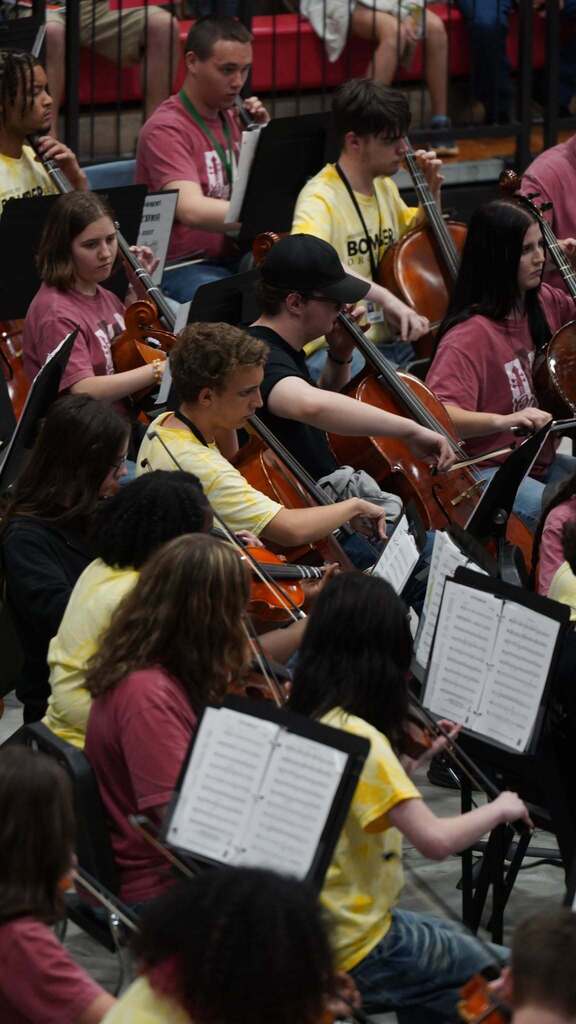 A group of people in a room play violins and sit on chairs. They are holding violins and bows.