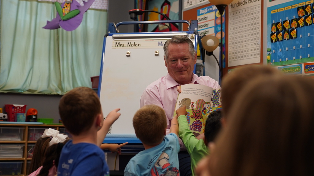 Man in pink shirt reading a book to a group of children, some of whom are raising their hands.