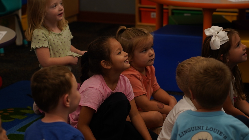 Group of young children in a circle, some sitting on the floor, facing forward in an indoor classroom setting.