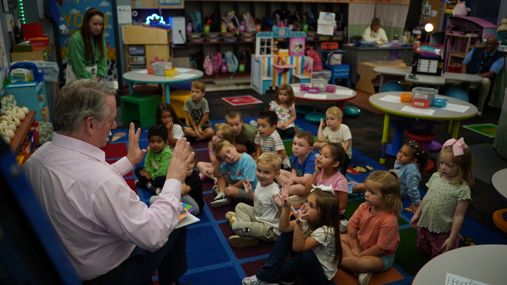 A man seated speaks to a group of children in a classroom. The children sit on a rug while some hold objects.