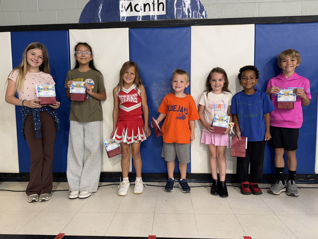 Eight young children pose for a photo holding bags while standing in front of a blue and white wall.