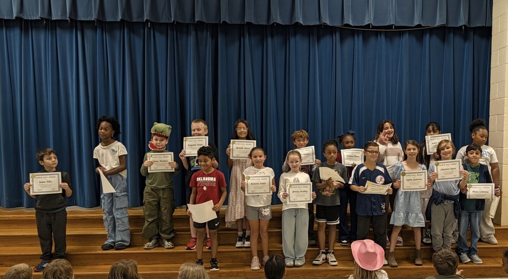 Group of students standing on stage holding certificates, blue curtain in background, some wearing hats.