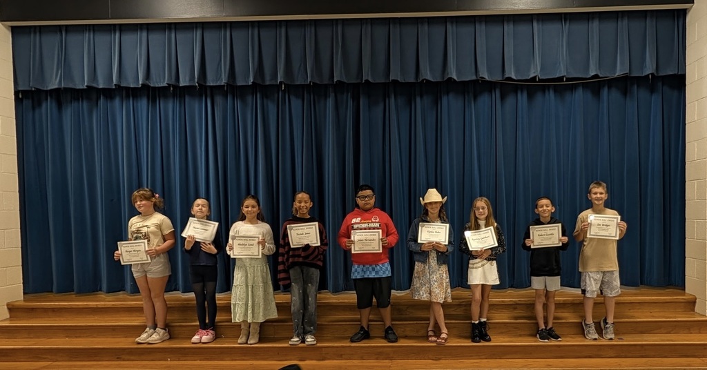 Students stand on stage, holding certificates, against a backdrop of a blue curtain.