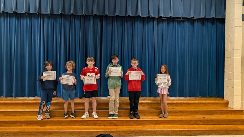 Seven children stand on a stage with wooden steps and blue curtains, holding certificates.