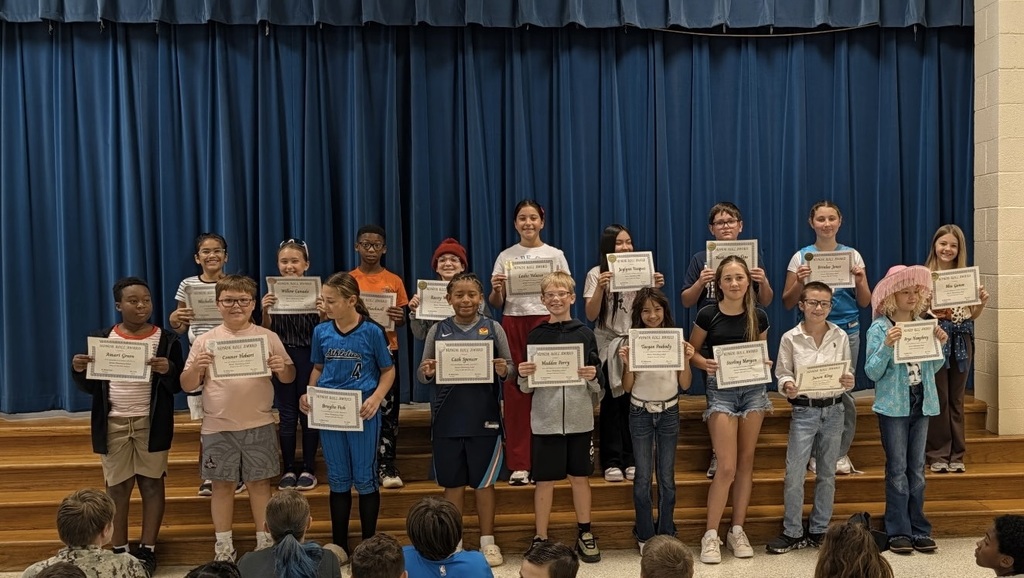 Children on a stage in front of a blue curtain, holding certificates and smiling at the camera.