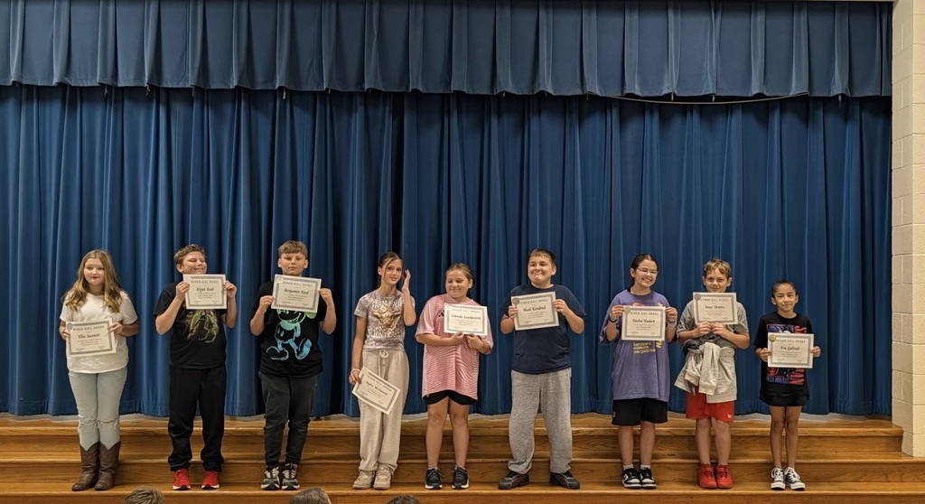 Students stand on stage, holding certificates, with a blue curtain in the background.