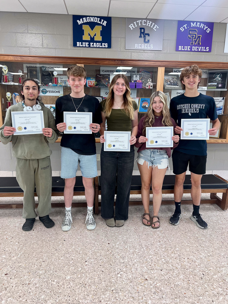 5 students smiling while holding certificates, standing in school lobby