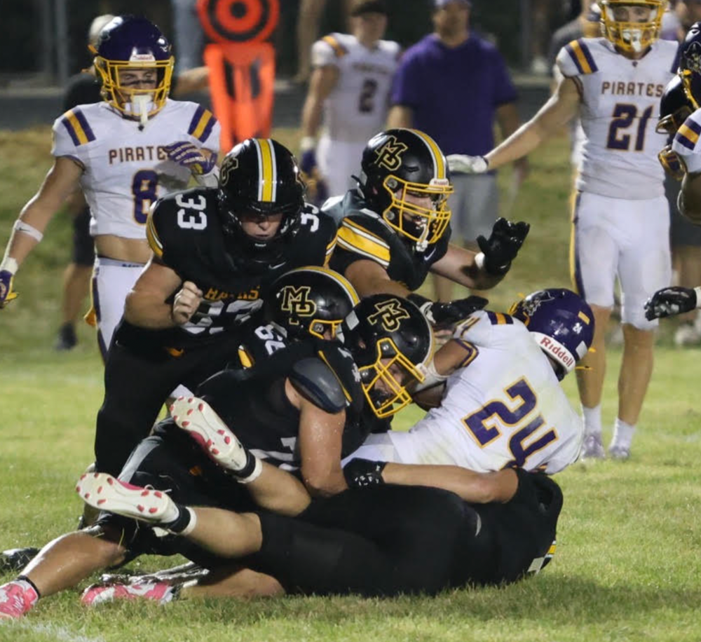 2 MP football players tackling another player in a white jersey on the football field