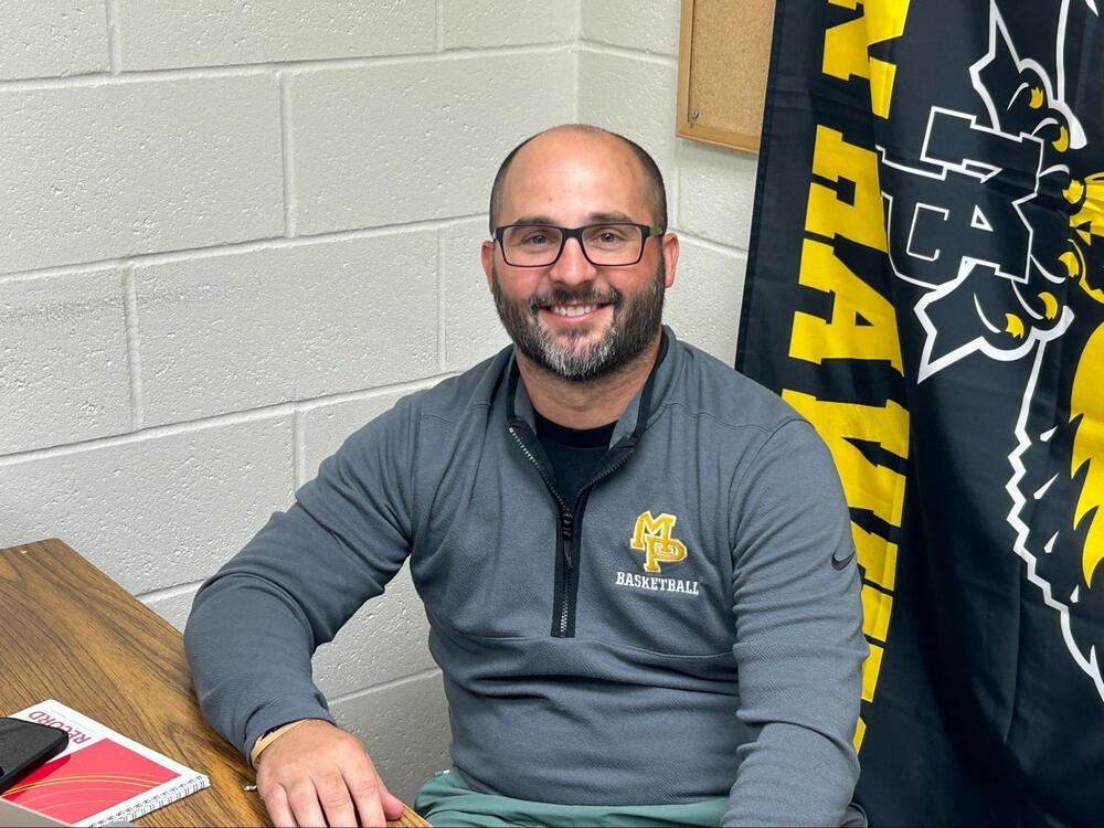 Justin Barthelman sitting at a desk wearing a grey quarter-zip shirt that says MP Basketball and a flag behind him that reads MP Hawks