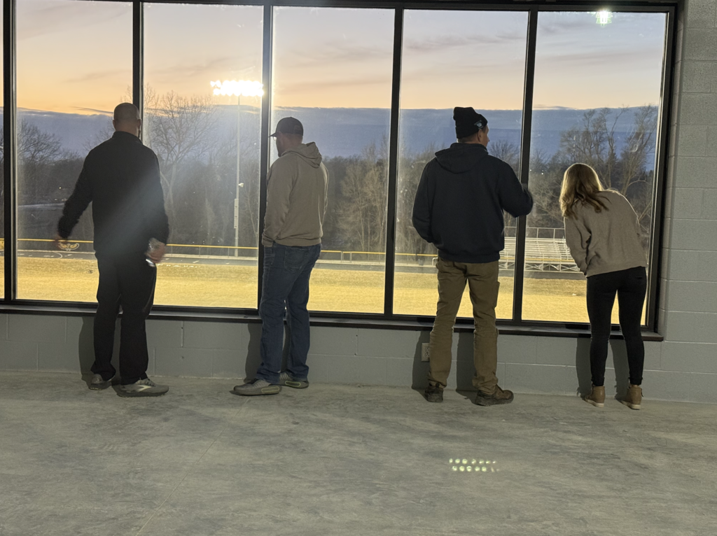 Facilities committee members standing at the large window of the activities building weight room overlooking the Mid-Prairie football field with a sunset in the background