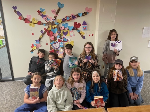 Elementary 3rd and 4th graders sitting in front of a Valentine's tree made out of hearts for leaves holding books